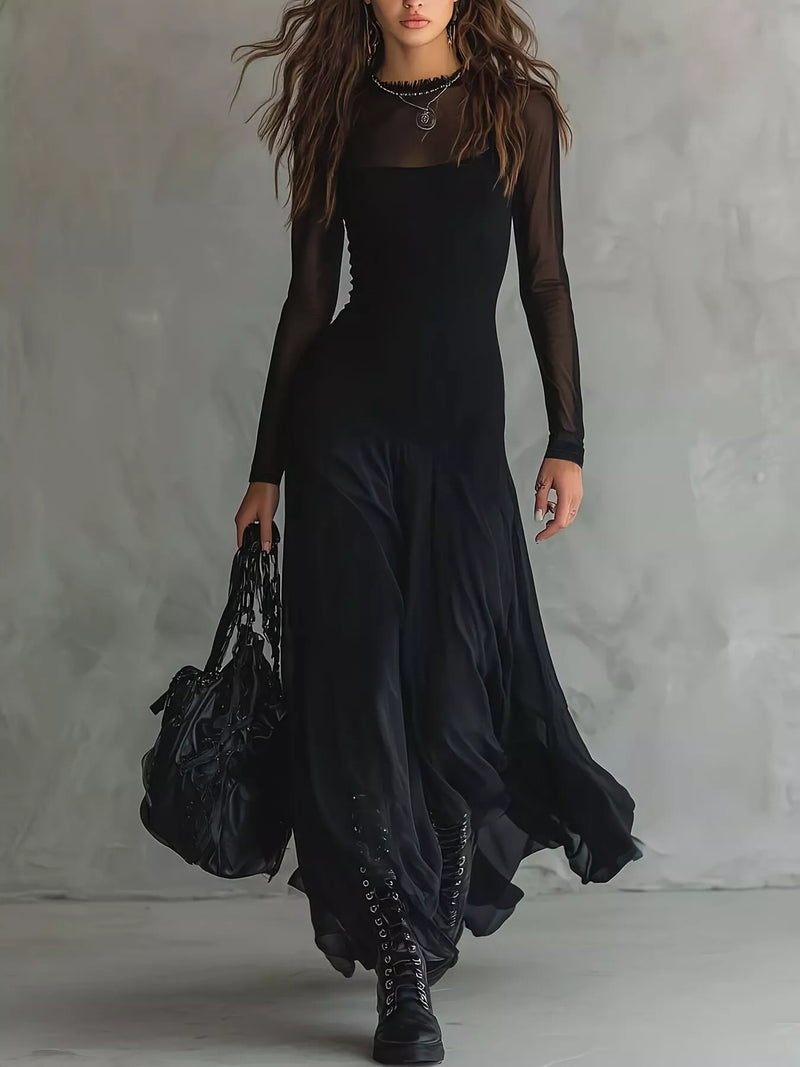 A woman models a long black dress with sheer long sleeves, a square neckline, and a flowing skirt, styled with black lace-up boots and a large black handbag against a textured gray studio backdrop.