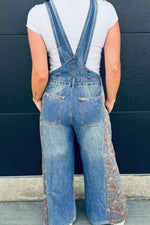 Person wearing floral-patterned denim overalls against a blue wall.