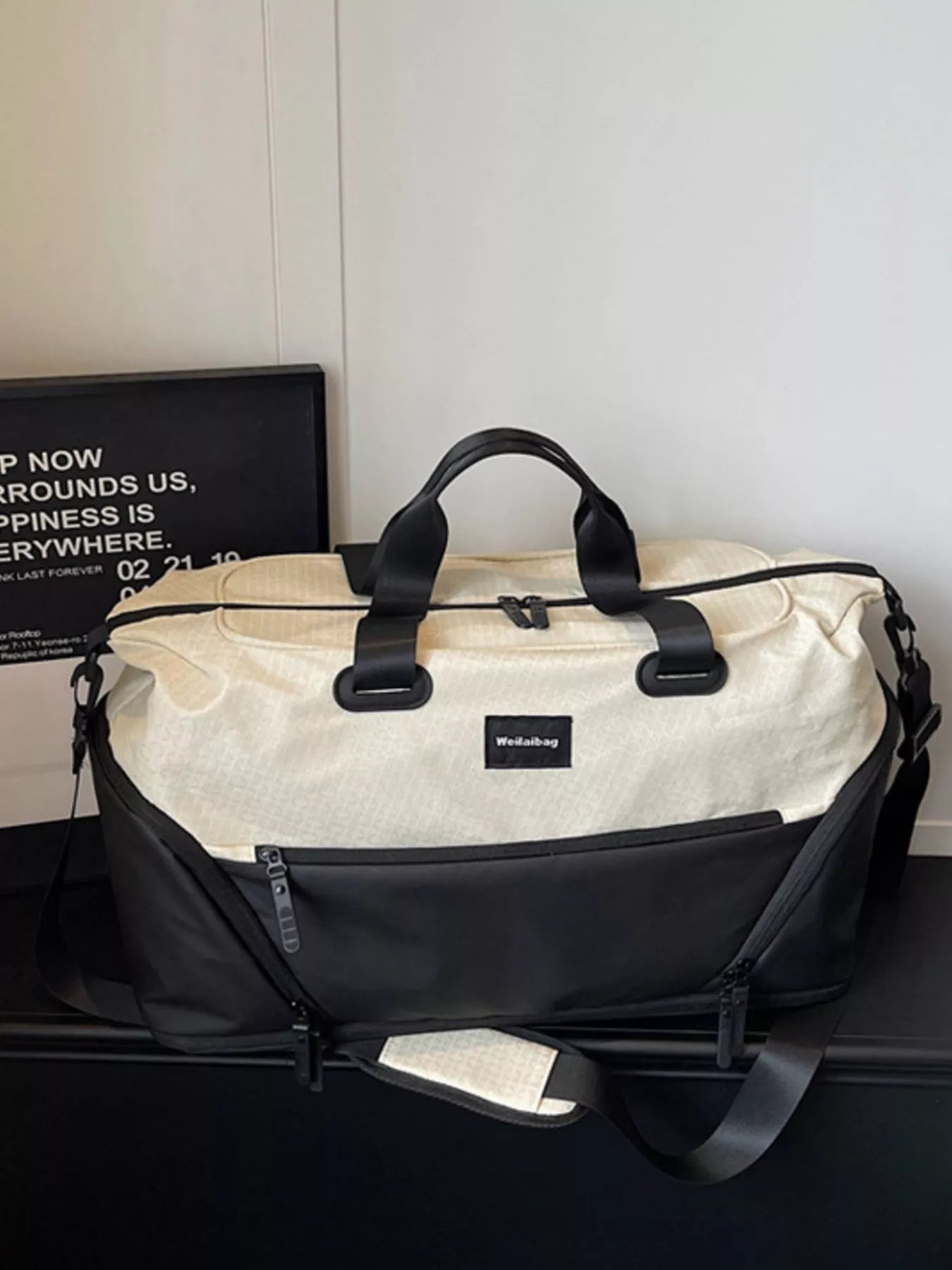 Beige and black duffel bag on a dark surface with a white wall background