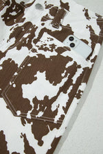 Close-up of brown and white cow-print denim jeans with a front pocket holding a white smartphone, photographed on a light textured surface.