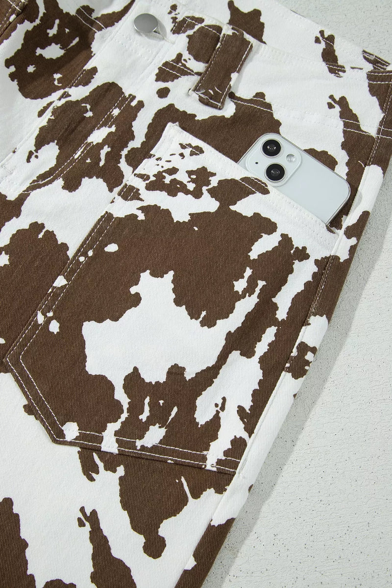 Close-up of brown and white cow-print denim jeans with a front pocket holding a white smartphone, photographed on a light textured surface.