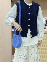 Person wearing a navy blue vest with gold buttons, holding a blue textured handbag.