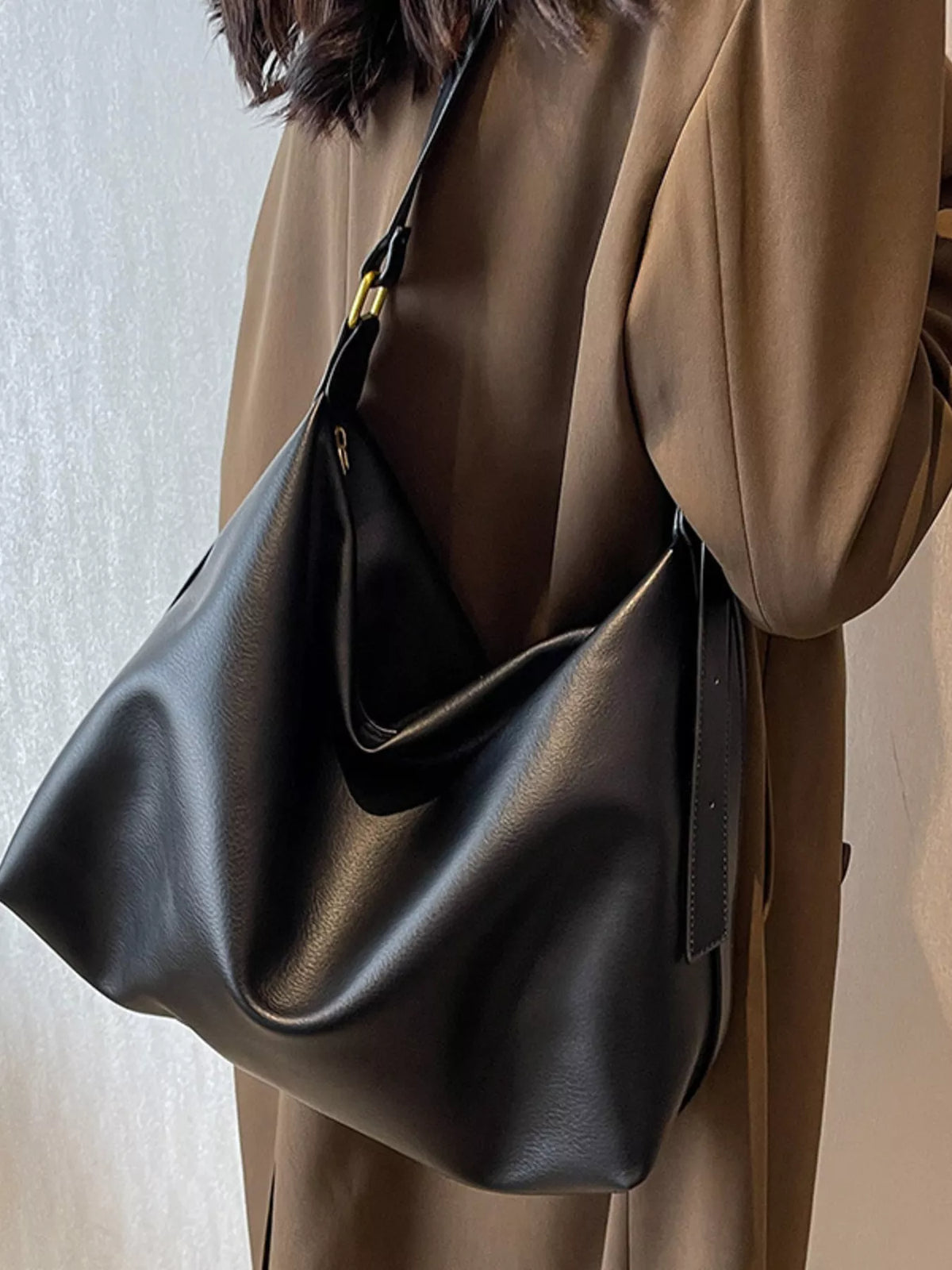 Person wearing a brown coat holding a black leather handbag against a neutral background