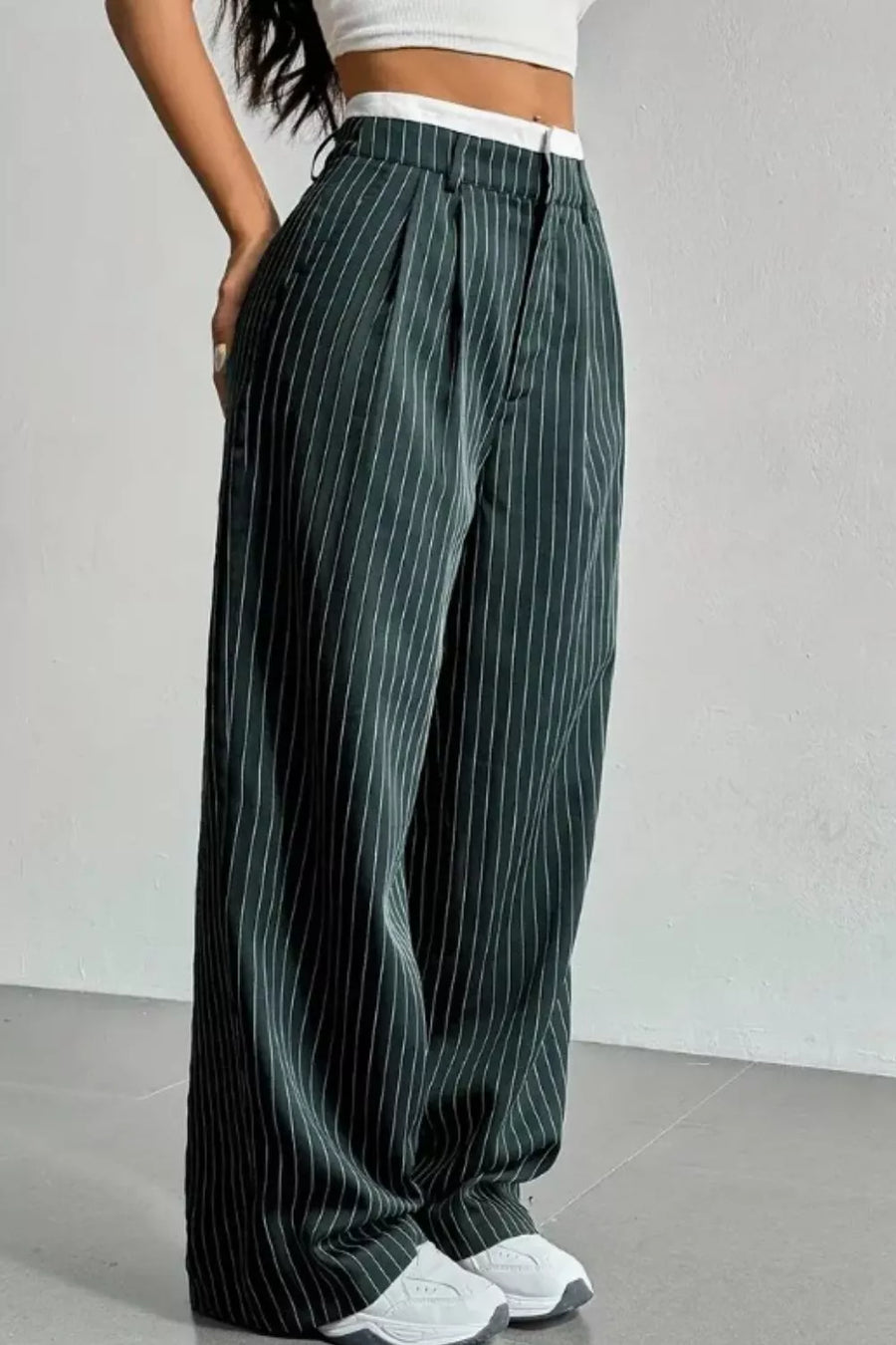 A person wearing dark green wide-leg pinstripe pants with a white waistband, paired with a white crop top and white sneakers, standing against a light gray wall.