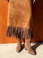 Person wearing a brown suede skirt with fringe and matching boots against an orange wall.