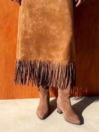 Person wearing a brown suede skirt with fringe and matching boots against an orange wall.