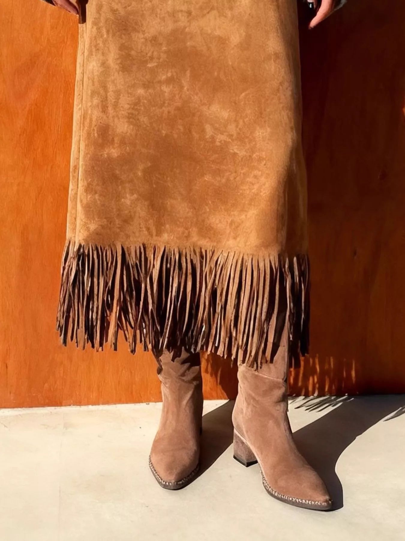 Person wearing a brown suede skirt with fringe and matching boots against an orange wall.