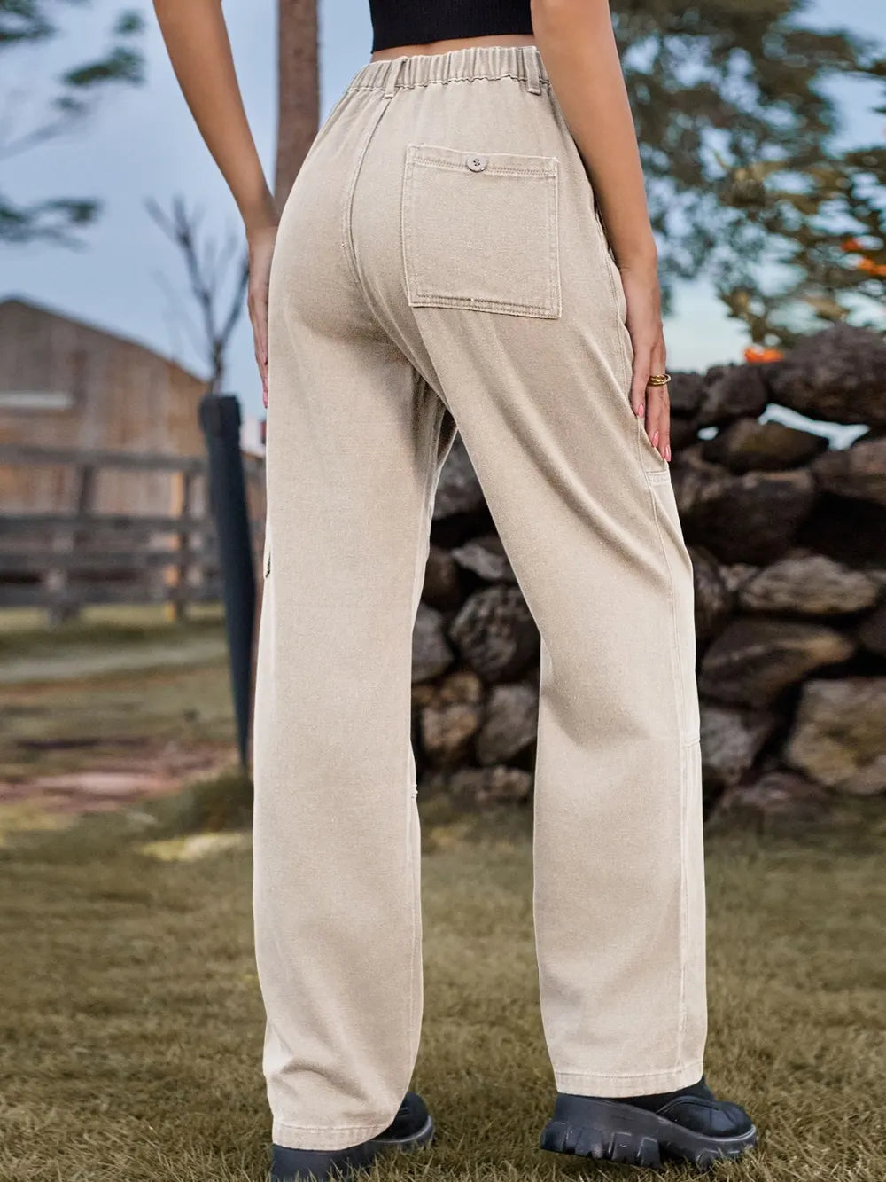 Person wearing beige pants standing outdoors with a wooden fence and stacked logs in the background
