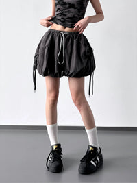 Person wearing a black skirt with drawstring, black top, white socks, and black shoes on a plain background