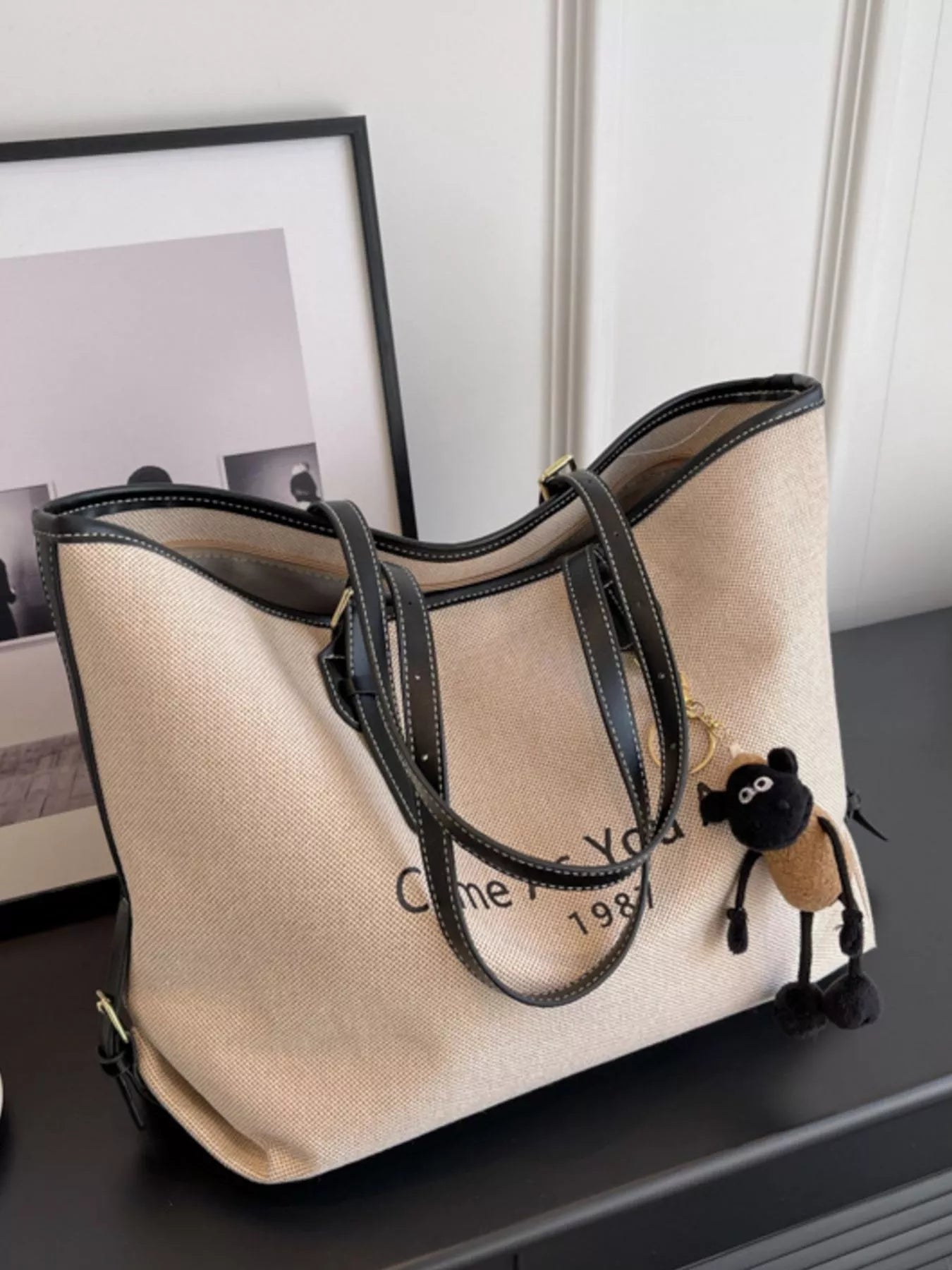 Beige tote bag with black handles and a small black and brown keychain on a dark surface.
