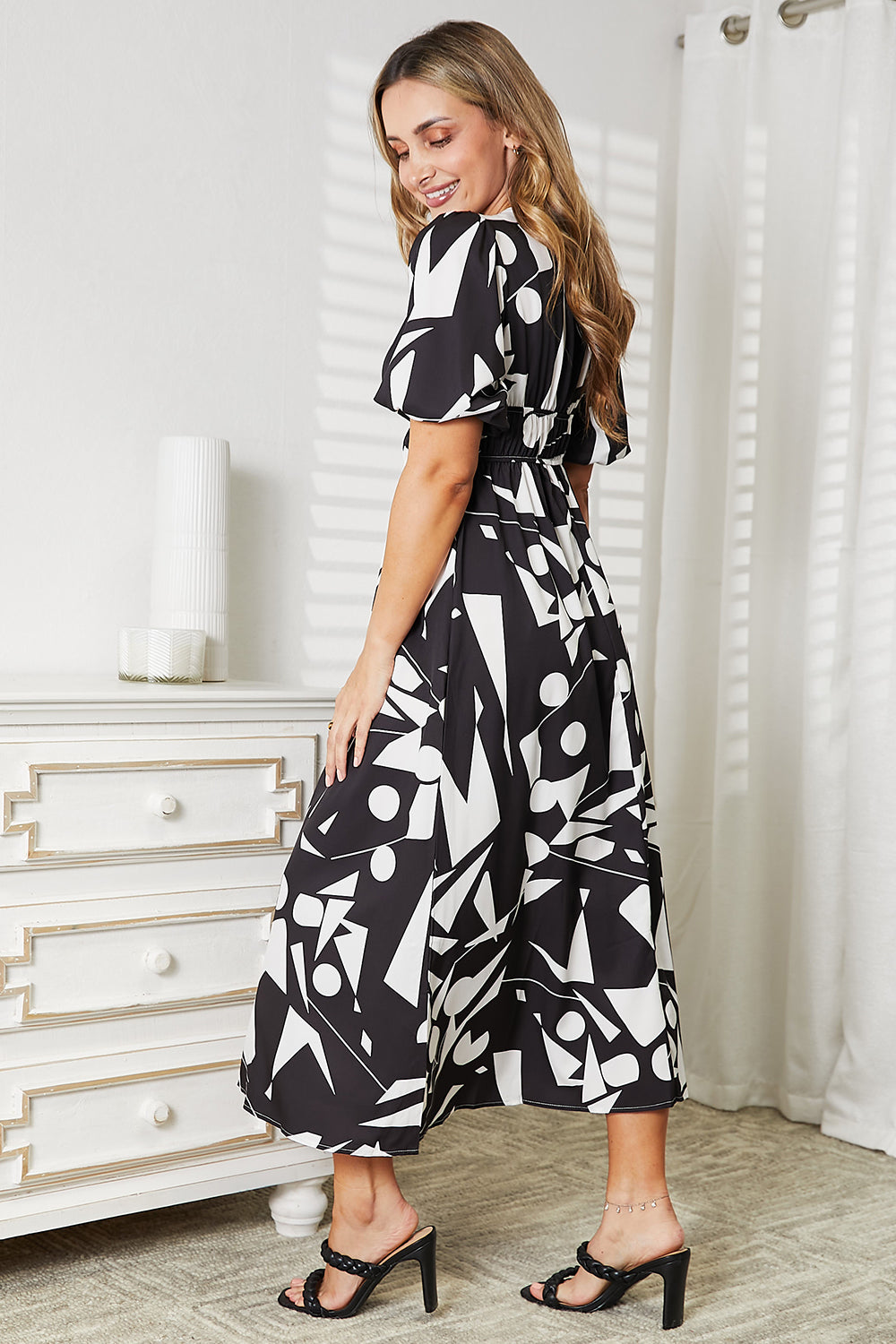 Woman wearing a black and white patterned dress in a room with a dresser and curtains.