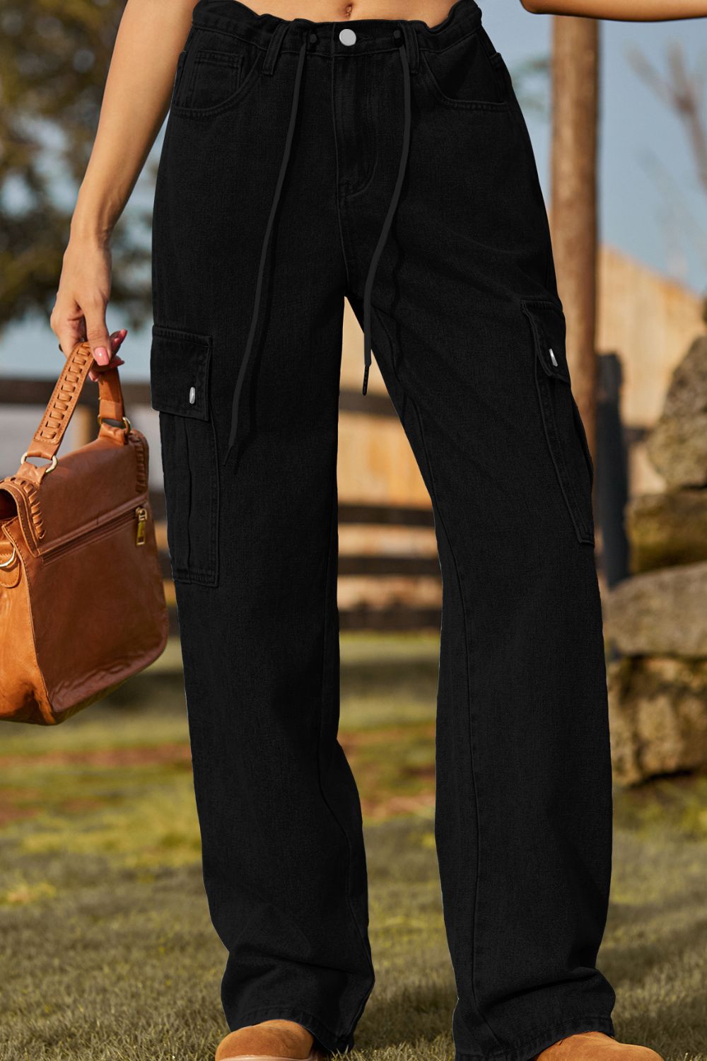 Person wearing black cargo pants holding a brown leather bag outdoors.