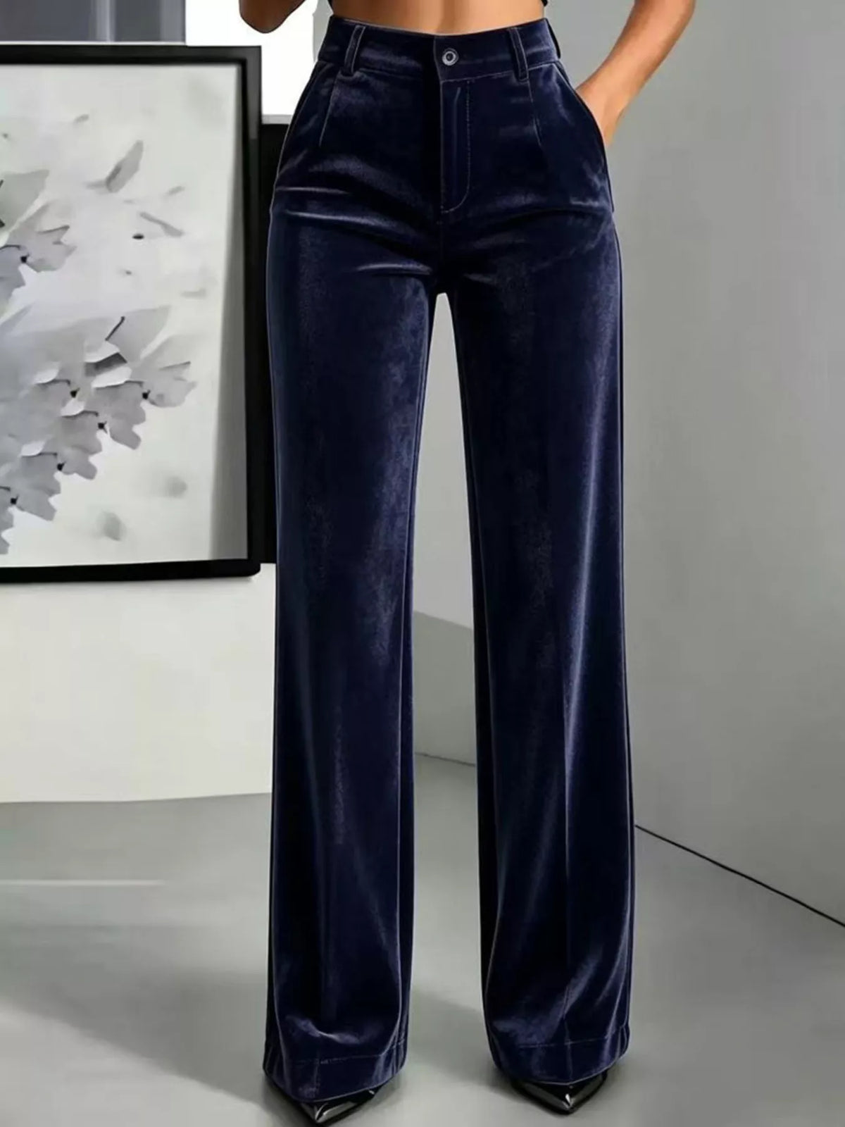 A person wearing high-waisted, wide-leg navy blue velvet pants, shown from the waist down in a modern indoor setting with a framed abstract print on the wall.