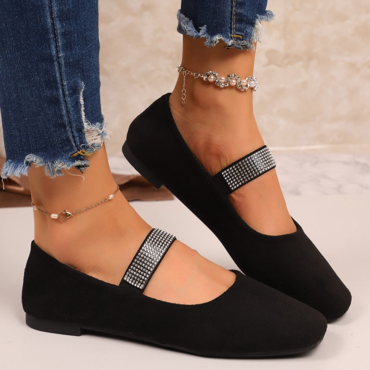 Black flat shoes with rhinestone straps worn with blue jeans on a neutral background