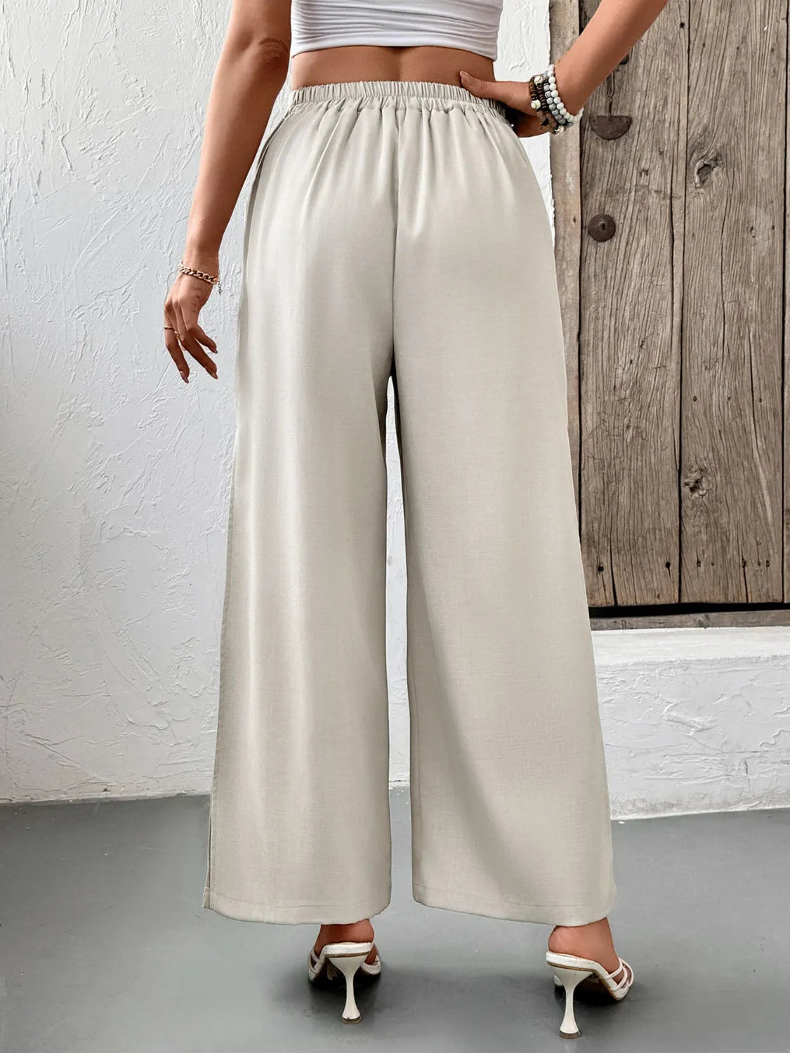 Person wearing beige wide-leg pants with a white top and high heels, standing against a wooden door.