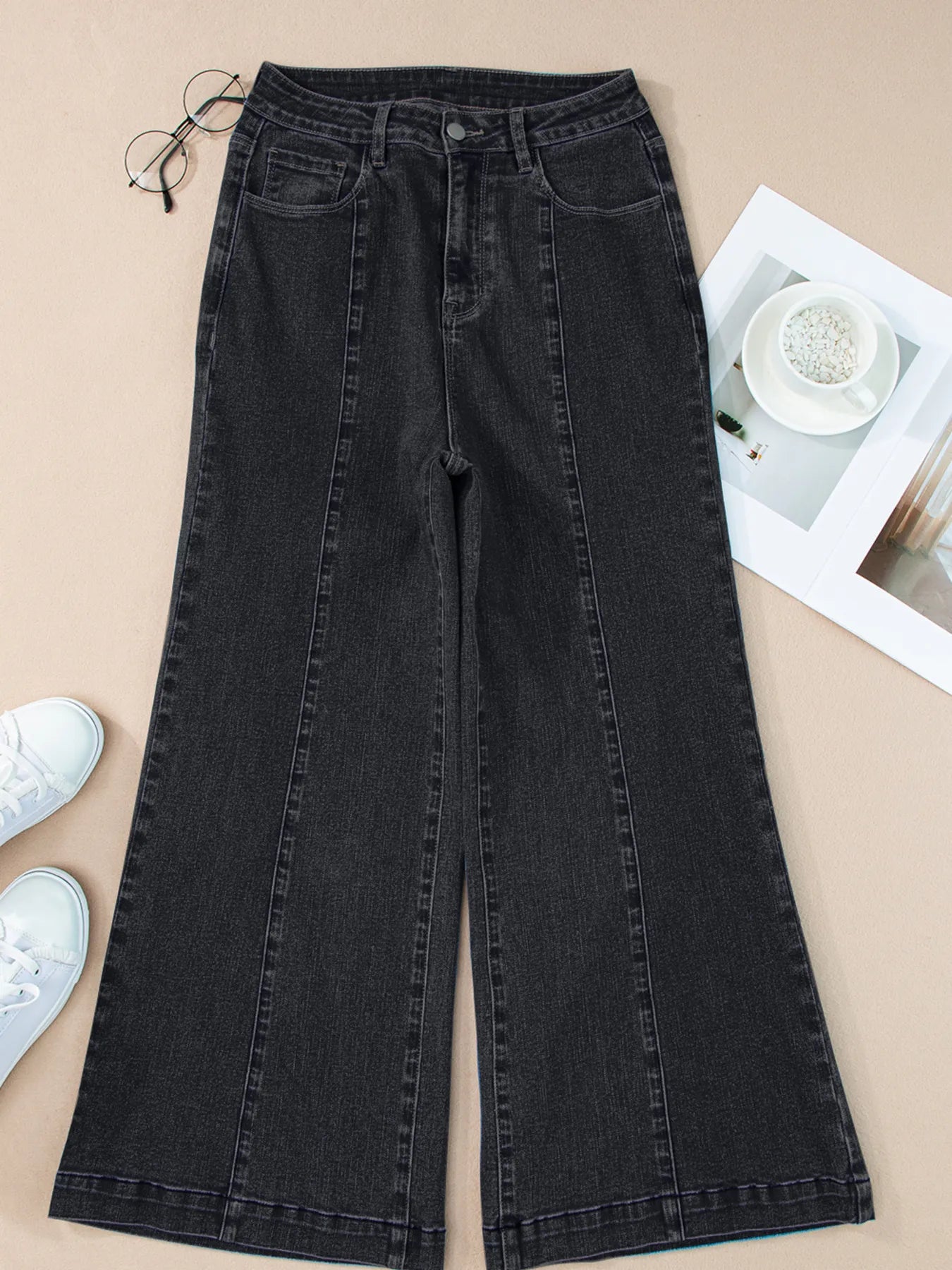 Black wide-leg jeans on a beige surface with white sneakers and eyeglasses.