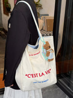 Person holding a tote bag with a teddy bear and text, standing indoors.