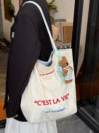Person holding a tote bag with a teddy bear and text, standing indoors.