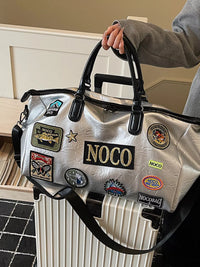 Silver bag with NOCO patches held by a person next to a suitcase.