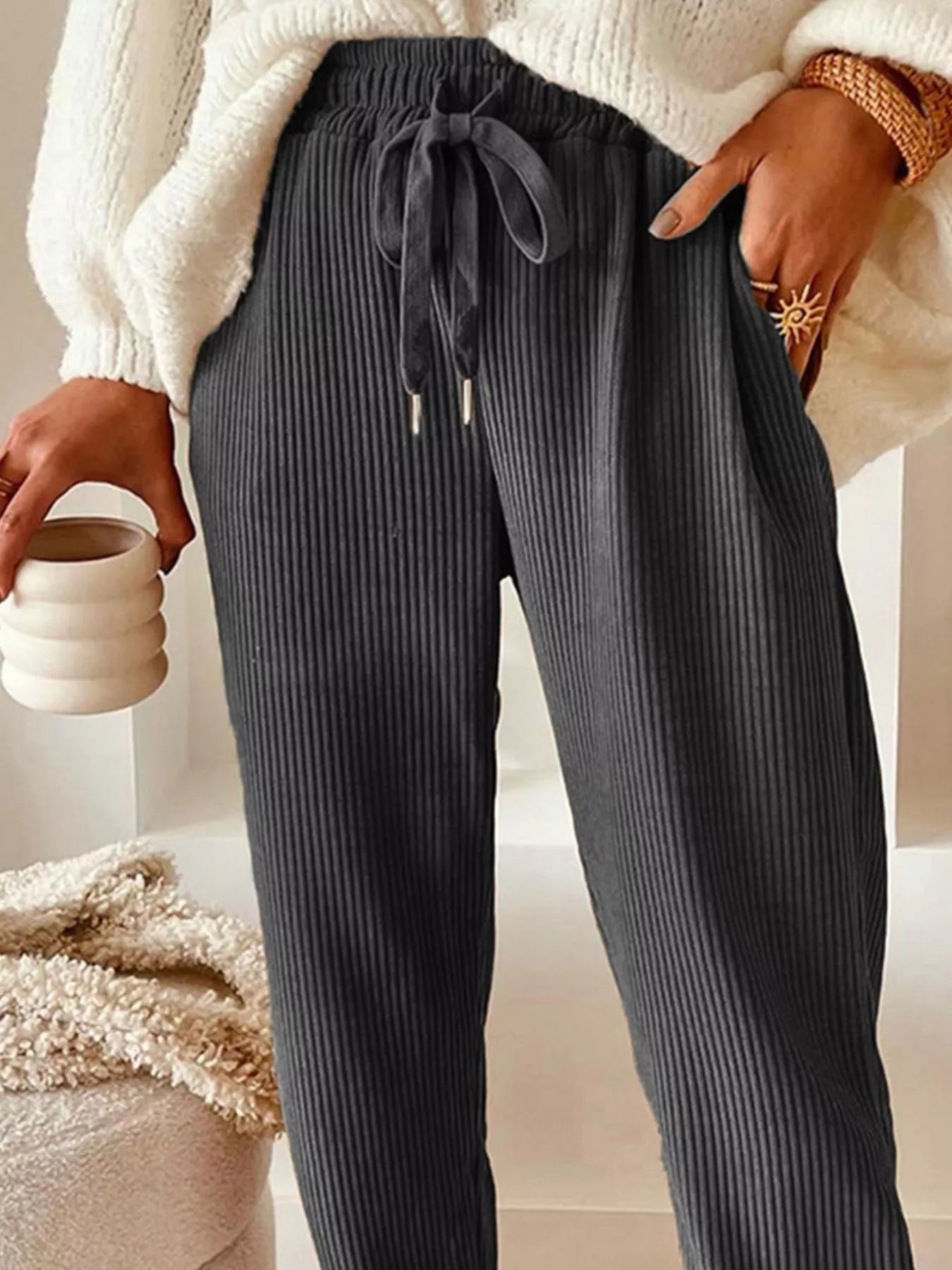 Person wearing dark ribbed pants with a white sweater and holding a ceramic cup.