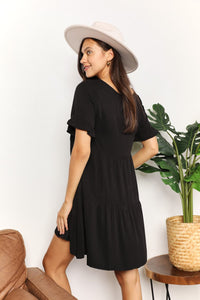 Woman wearing a black dress and beige hat standing next to a plant indoors.