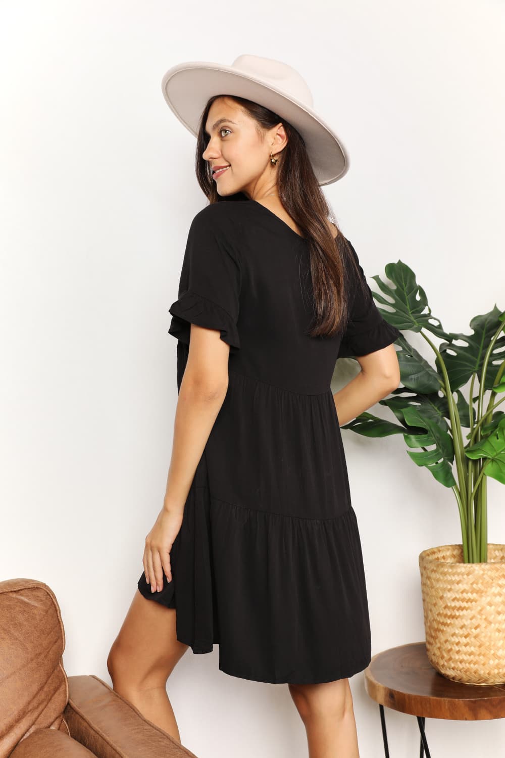 Woman wearing a black dress and beige hat standing next to a plant indoors.