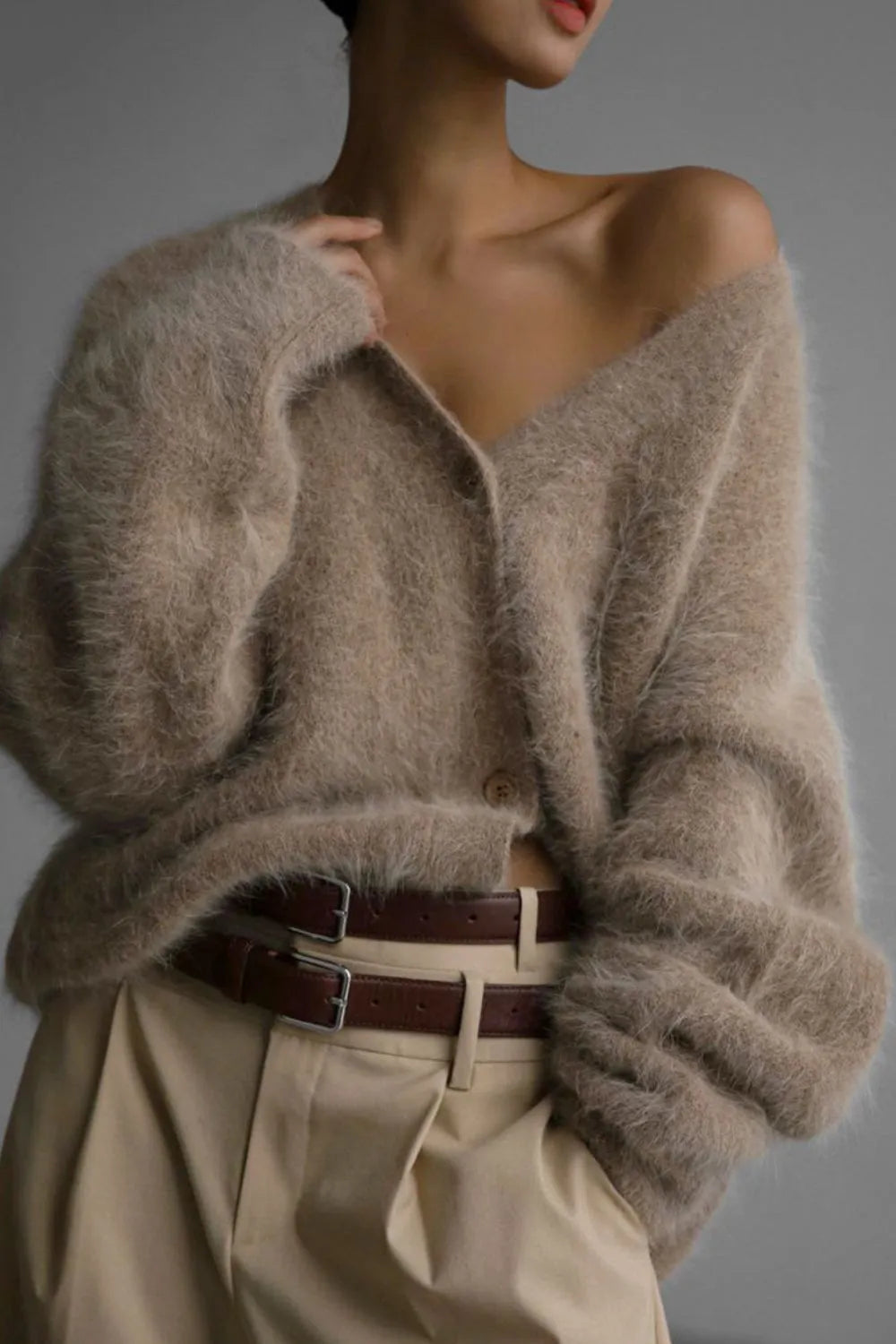 Person wearing a beige cardigan and beige pants with a brown belt against a gray background