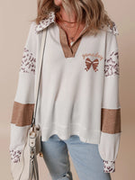 Person wearing a white sweater with leopard print sleeves and a brown bow design, holding a beige handbag.