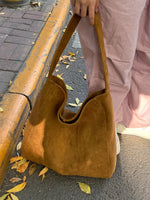 A person wearing dusty pink trousers holds a large, slouchy brown suede shoulder bag on an outdoor paved street with scattered autumn leaves.