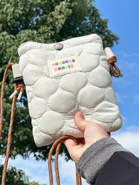 White quilted pouch with colorful label held by a hand against a blue sky.
