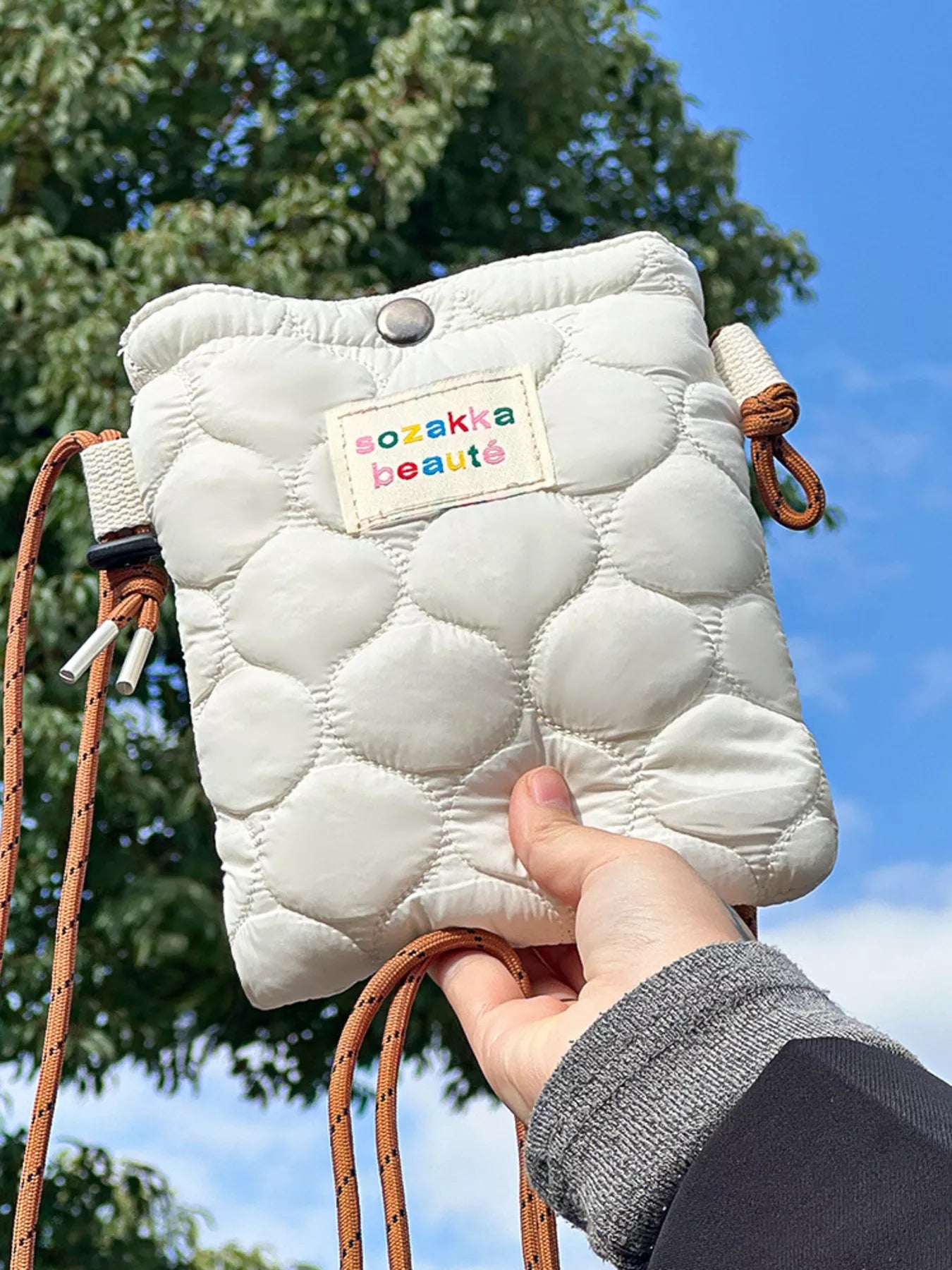 White quilted pouch with colorful label held by a hand against a blue sky.