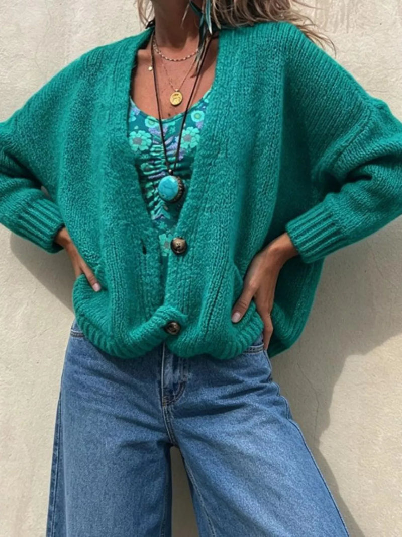 A woman wearing a chunky knit green cardigan over a teal floral top and blue jeans, standing against a textured beige wall.