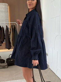 Woman wearing a navy blue dress in a room with a coat rack and bags in the background