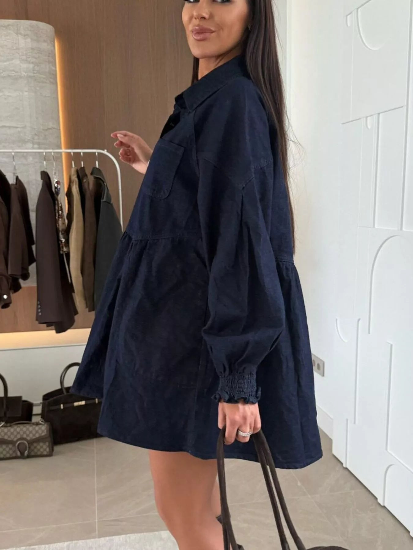 Woman wearing a navy blue dress in a room with a coat rack and bags in the background