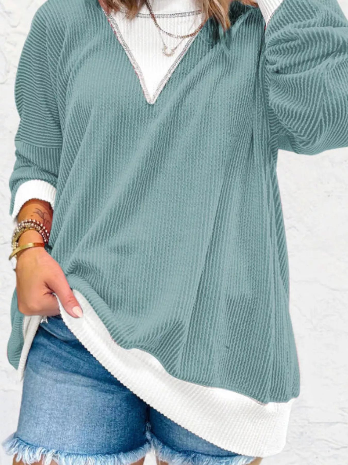 Person wearing a teal sweater over a white shirt with denim shorts on a white background