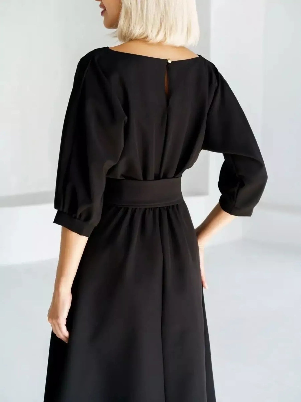A woman wearing a black midi dress with three-quarter puff sleeves and a matching fabric belt at the waist, shown from the back in a bright, minimalist studio setting.