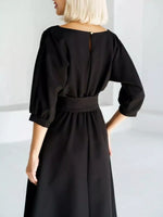 A woman wearing a black midi dress with three-quarter puff sleeves and a matching fabric belt at the waist, shown from the back in a bright, minimalist studio setting.