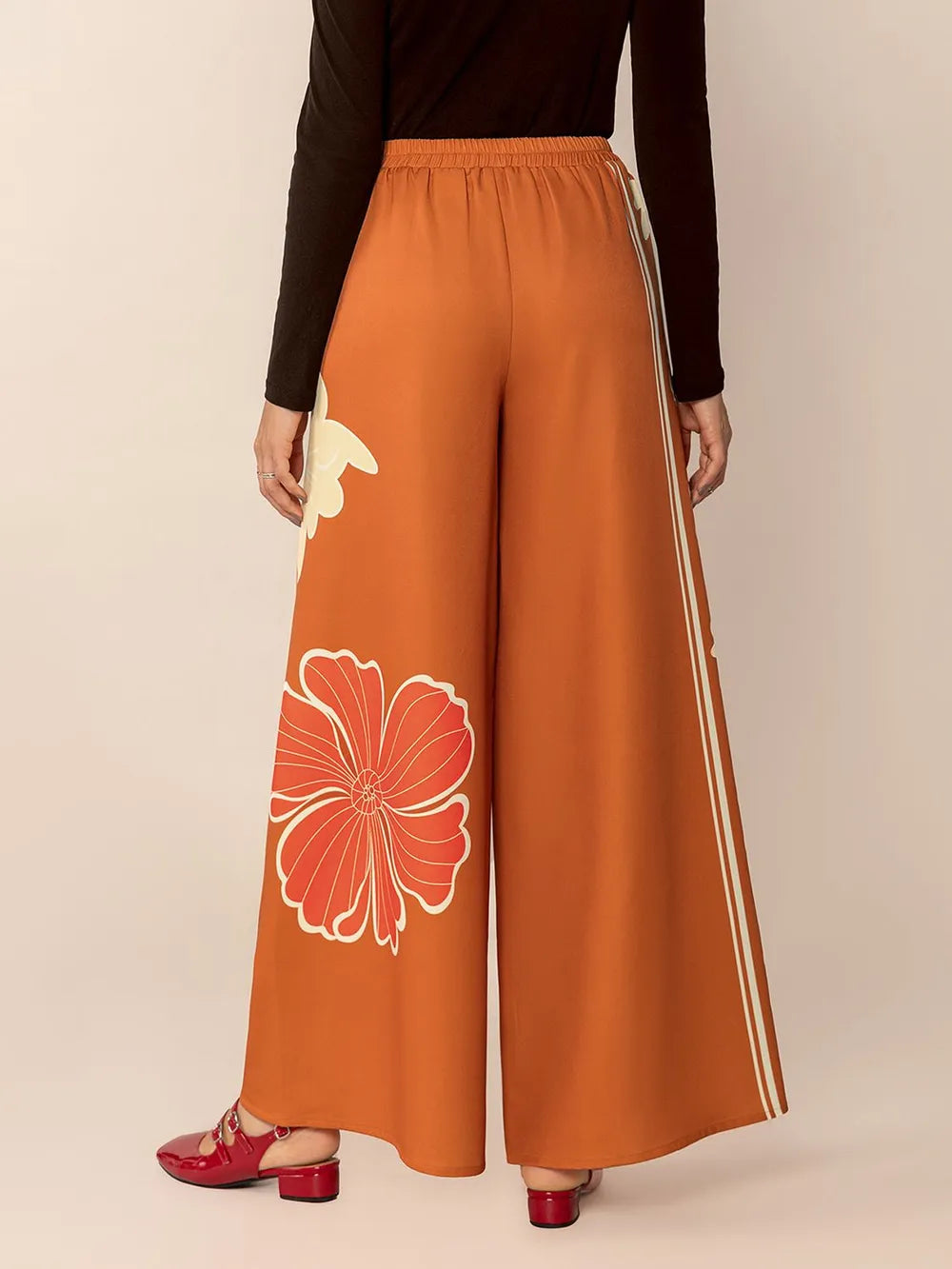 Orange wide-leg pants with floral design and white stripes worn by a person on a beige background