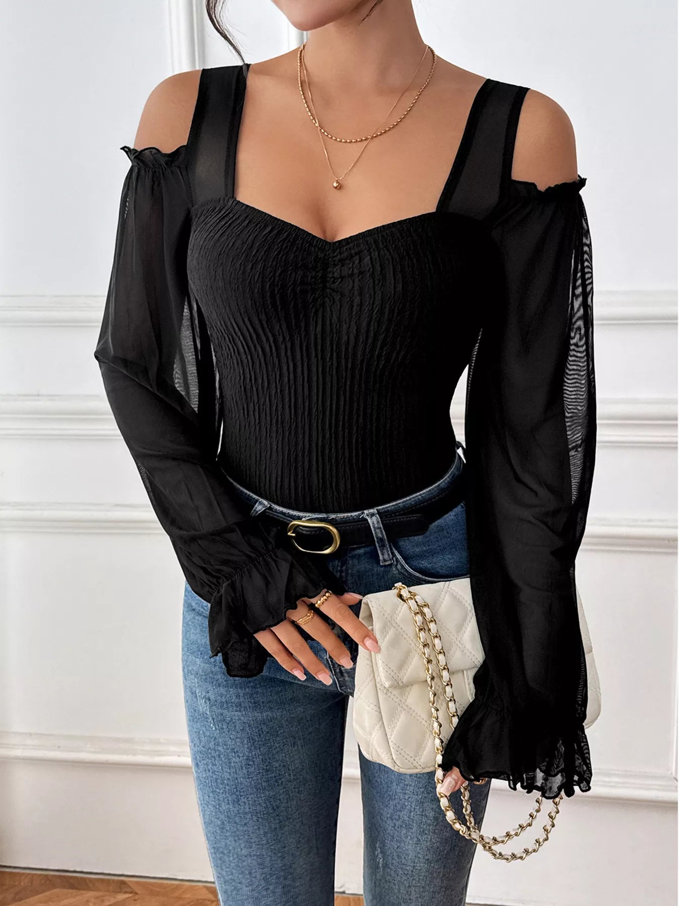 Woman wearing a black off-shoulder top with sheer sleeves and blue jeans, holding a white handbag.