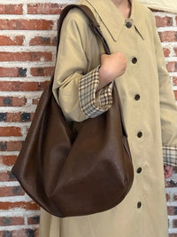 Person wearing a beige coat holding a brown leather bag against a brick wall.