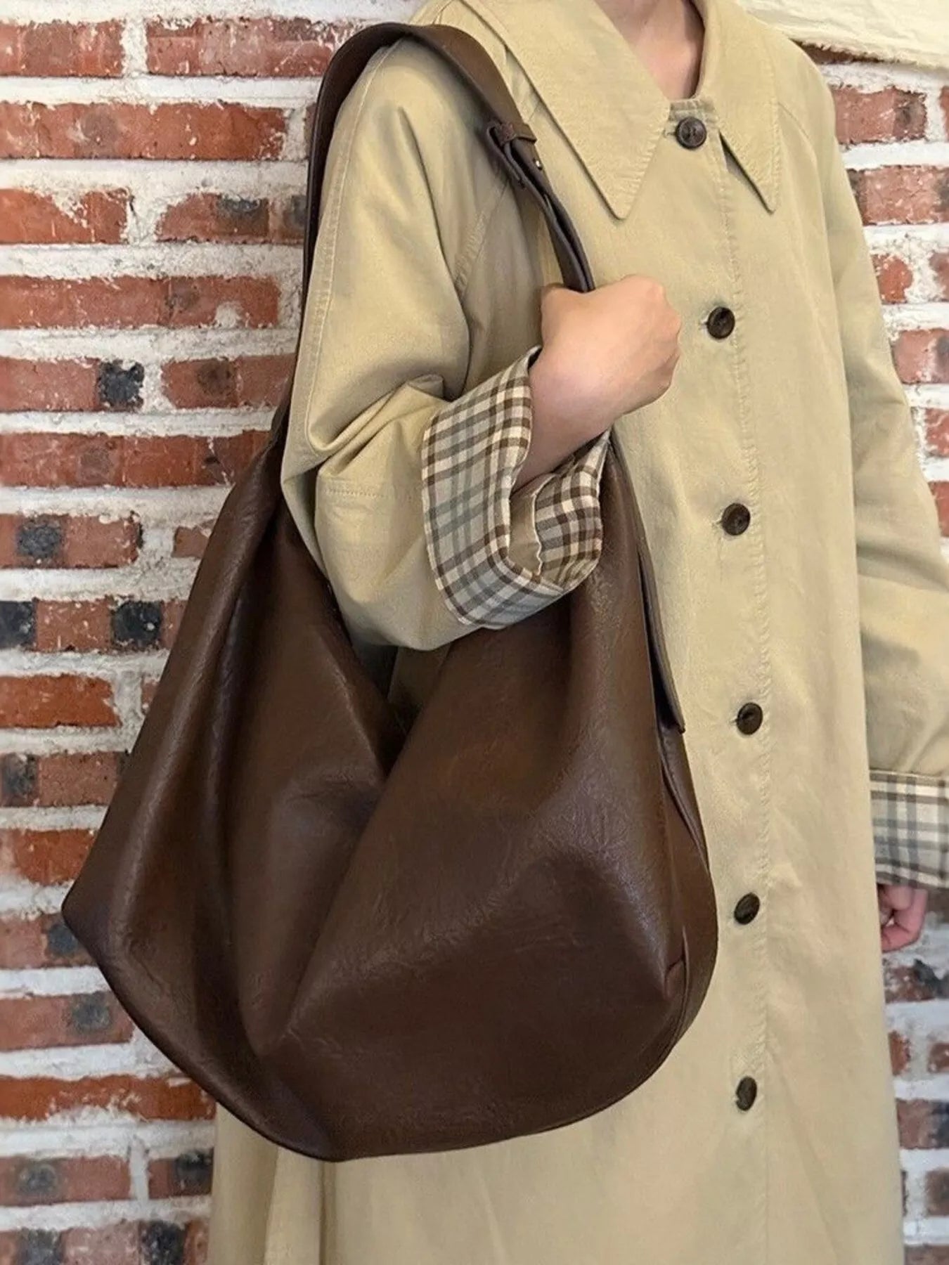 Person wearing a beige coat holding a brown leather bag against a brick wall.
