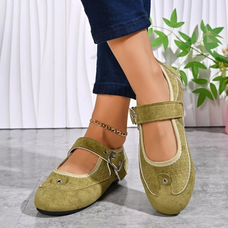 A person wearing olive green Mary Jane style flat shoes with decorative metal eyelets and buckle straps, paired with dark blue jeans. The shoes are shown on a gray floor against a white curtain background with a green plant visible.