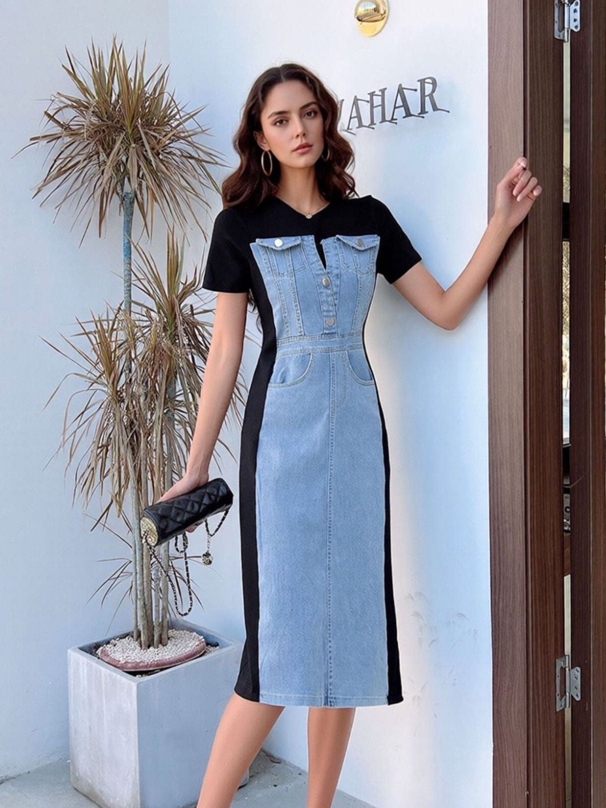 Woman wearing a black and denim dress standing against a light blue wall with a plant and door in the background.
