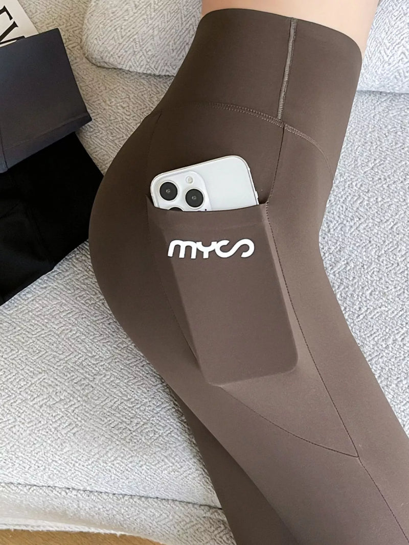 A close-up of a person's leg wearing brown MYCO ankle-length leggings, showing a side pocket holding a white smartphone. The background is a textured light gray sofa.