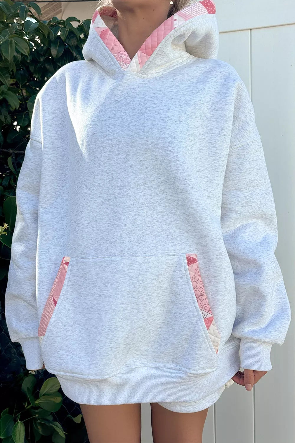 A person wearing an oversized light heather gray hoodie with pink patterned lining details on the hood and front pocket openings, photographed outdoors against a white door and greenery.