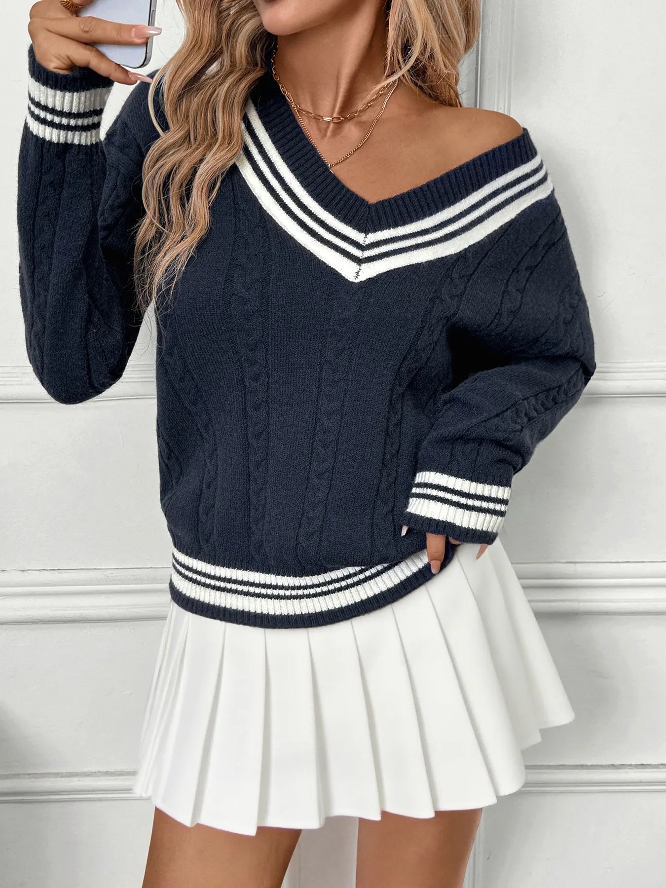Woman wearing a navy blue sweater with white stripes and a white pleated skirt against a white wall.