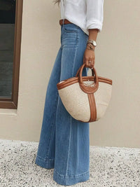 Person holding a straw handbag with brown handles wearing blue jeans and a white shirt.