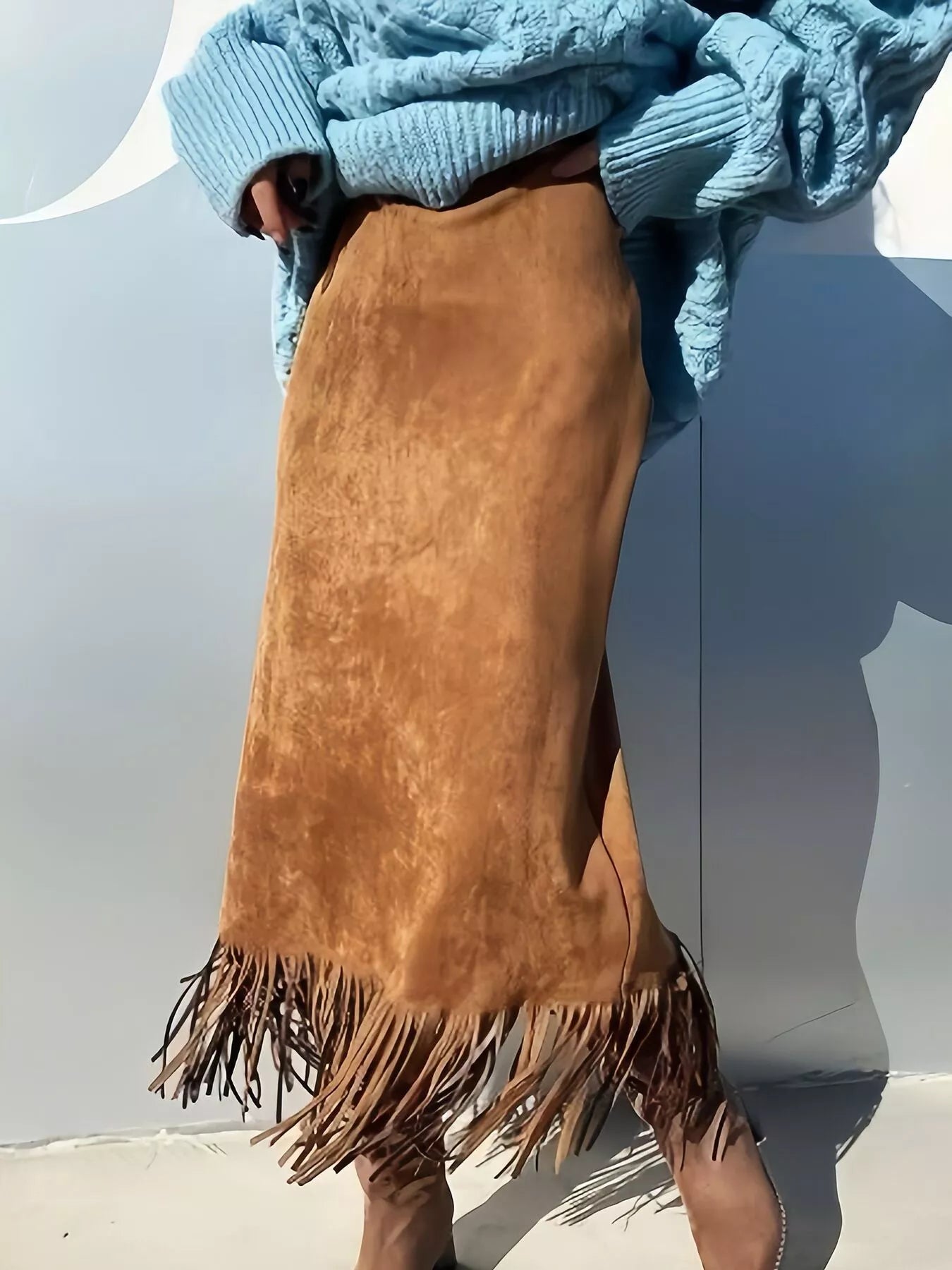 Person wearing a brown suede skirt with fringe and a blue sweater against a light gray background