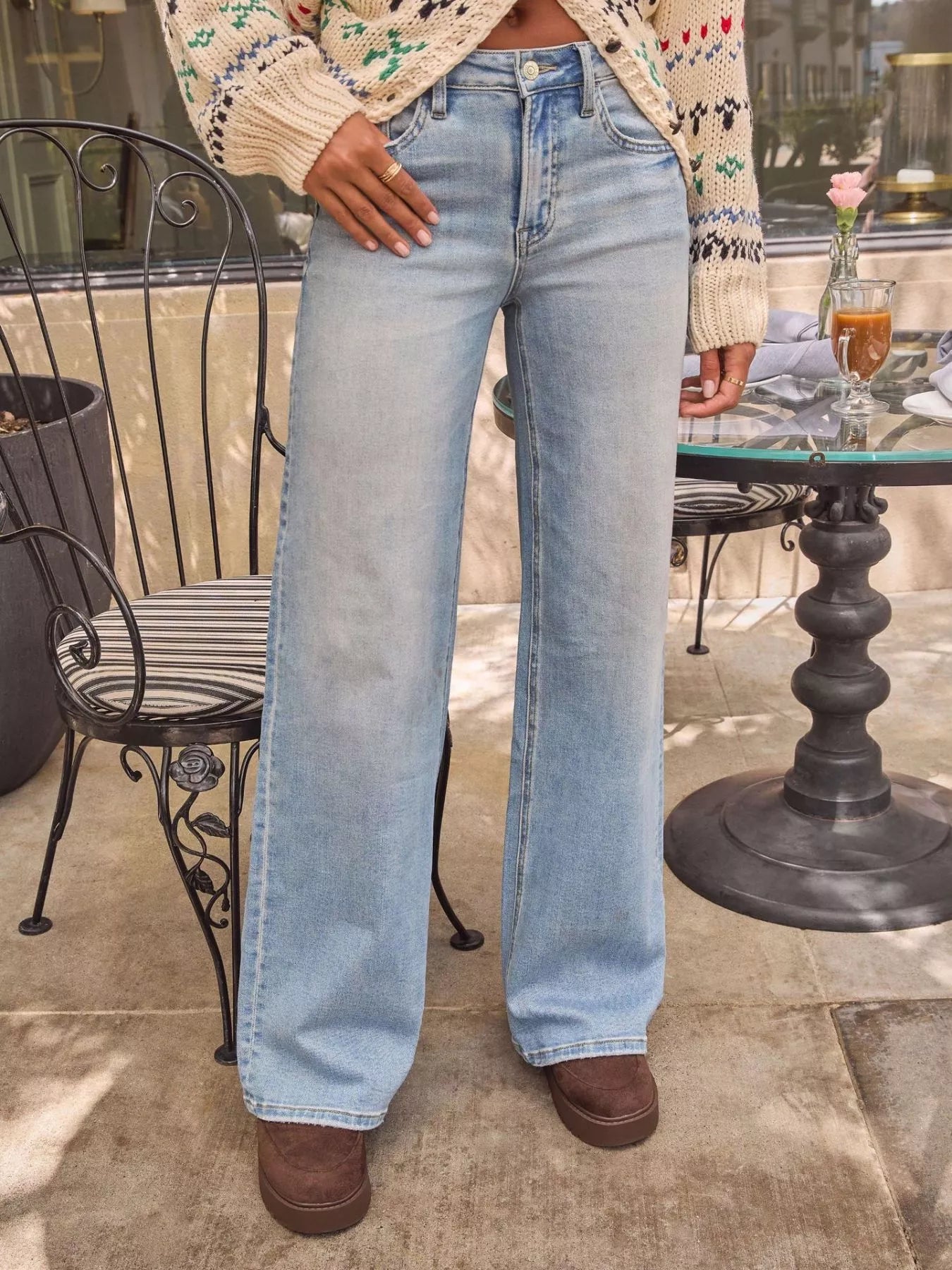 Person wearing light blue jeans standing outdoors near a table with a plant and drink.