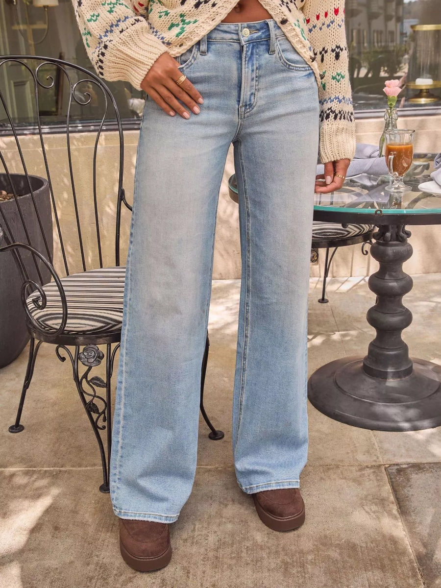 Person wearing light blue jeans standing outdoors near a table with a plant and drink.